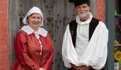 A woman and a man in seventeenth century clothes standing in front of a large, old wooden door.